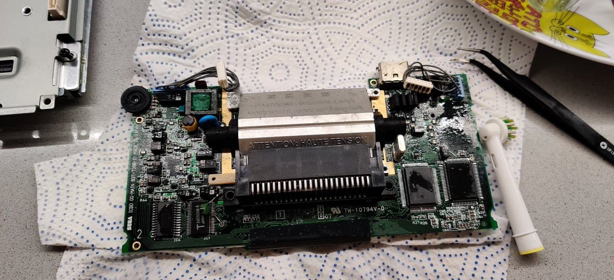 Disassembled Game Gear main PCB showing capacitor damage, corroded solder joints, and electrolyte residue with cleaning tools nearby.