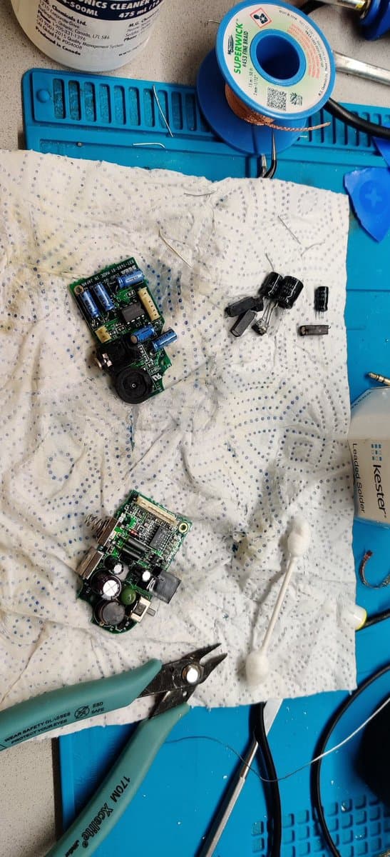 Desoldered Game Gear components on anti-static mat: two PCBs, electrolytic capacitors, ICs, and transistors; tweezers and solder spool visible above.