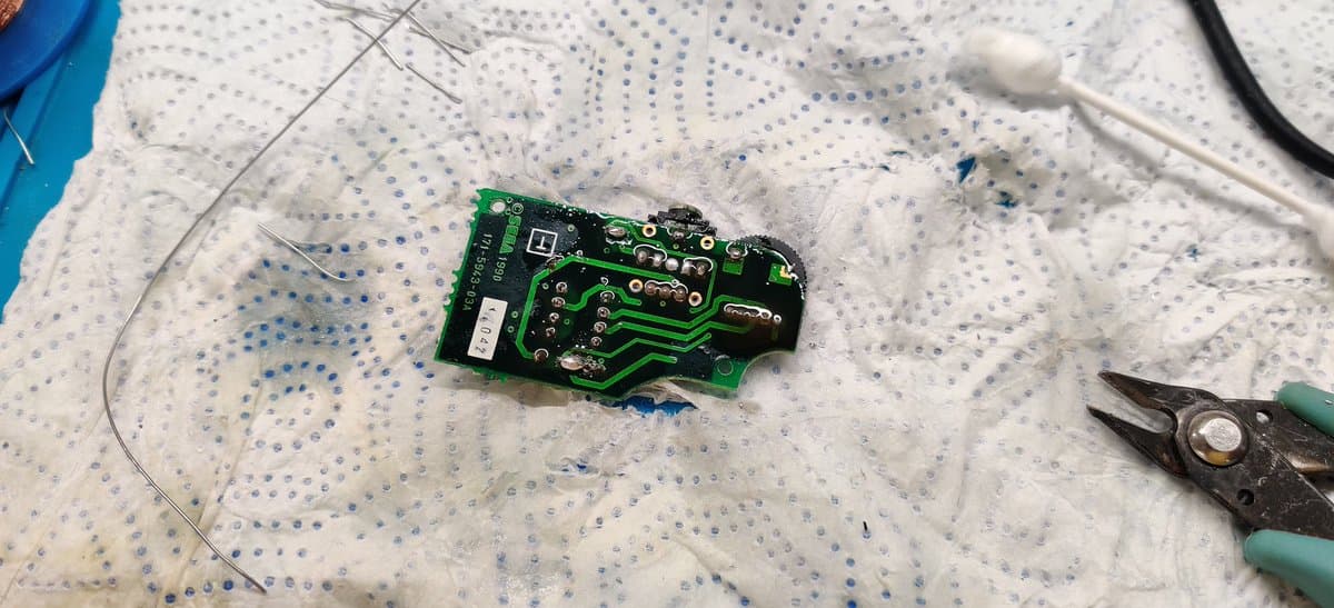 Green circuit board with exposed solder joints and traces, damaged components, flux residue, tweezers and wire nearby on work mat during Game Gear capacitor leak repair.