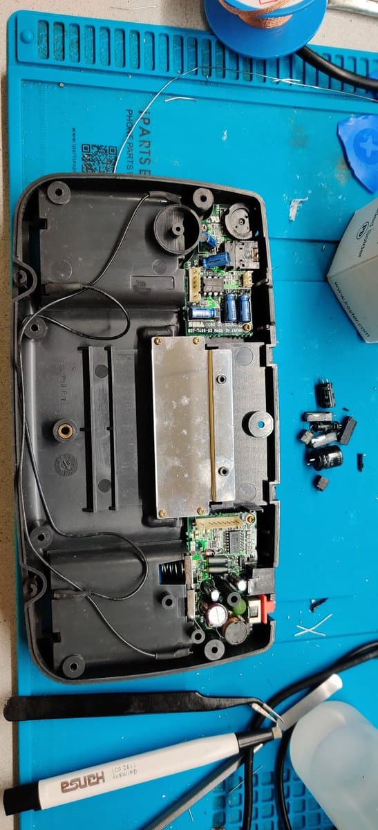 Disassembled Game Gear interior showing corroded PCB with leaking capacitor damage, two circuit boards, and separated components on blue workmat with soldering iron.