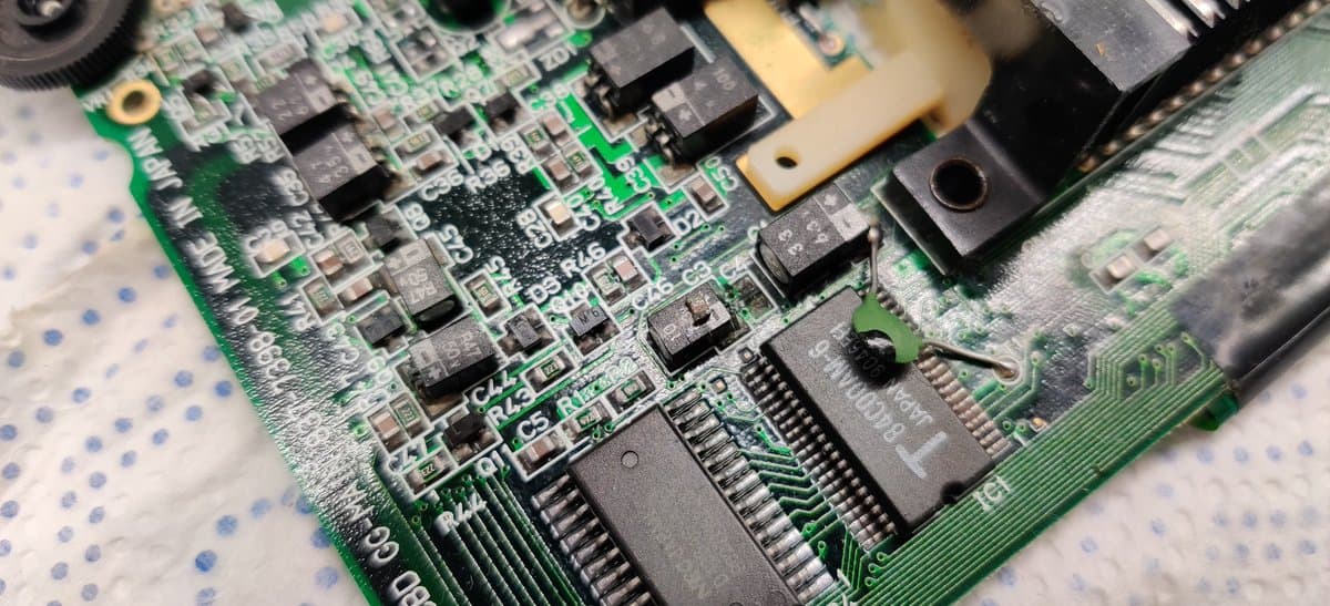 Heavily corroded Game Gear PCB with electrolyte damage; ICs and resistors partially detached from pads, green board visible beneath contamination.