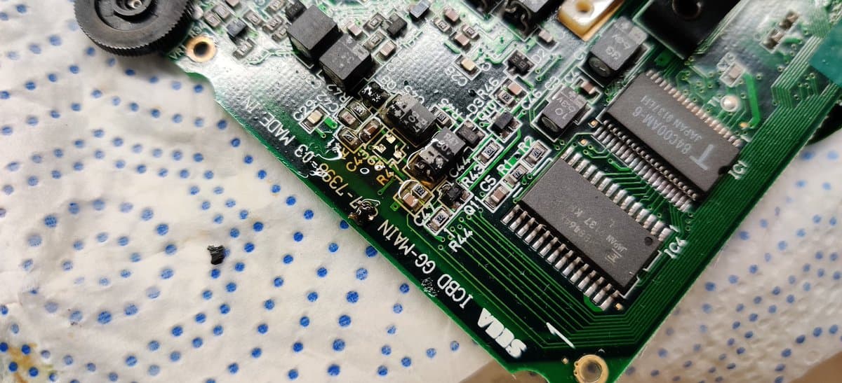 Sega Game Gear PCB with electrolyte damage; ICs and discrete components detached from corroded pads on green circuit board.