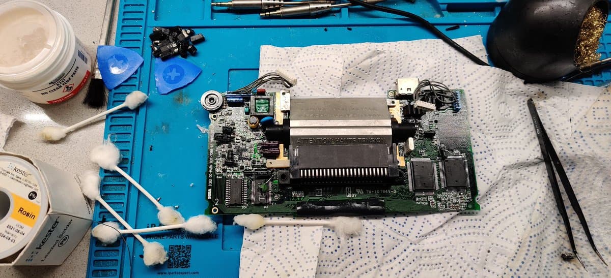 Two Sega Game Gear main PCBs on blue mat with cleaning supplies, cotton swabs with dried electrolyte residue, partially cleaned boards showing component damage and corrosion repair work completed.