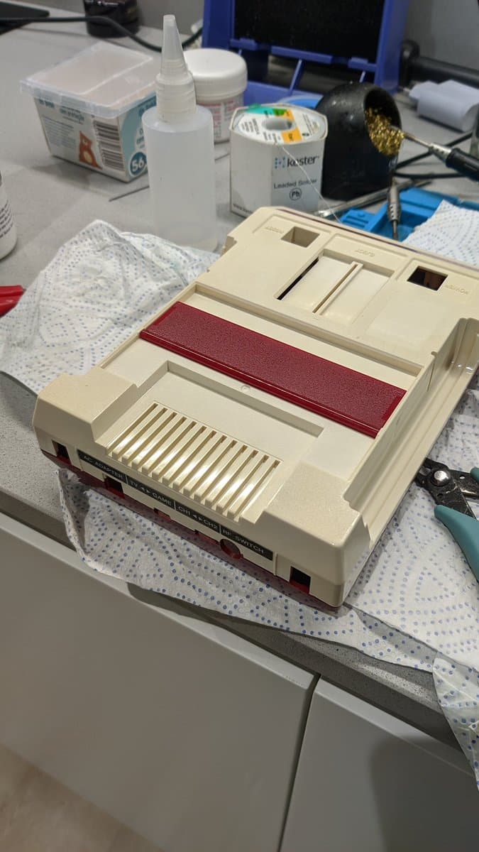 Repaired Famicom with composite mod board installed, burgundy heat shrink visible, sitting on work mat surrounded by solder, flux, and repair supplies.