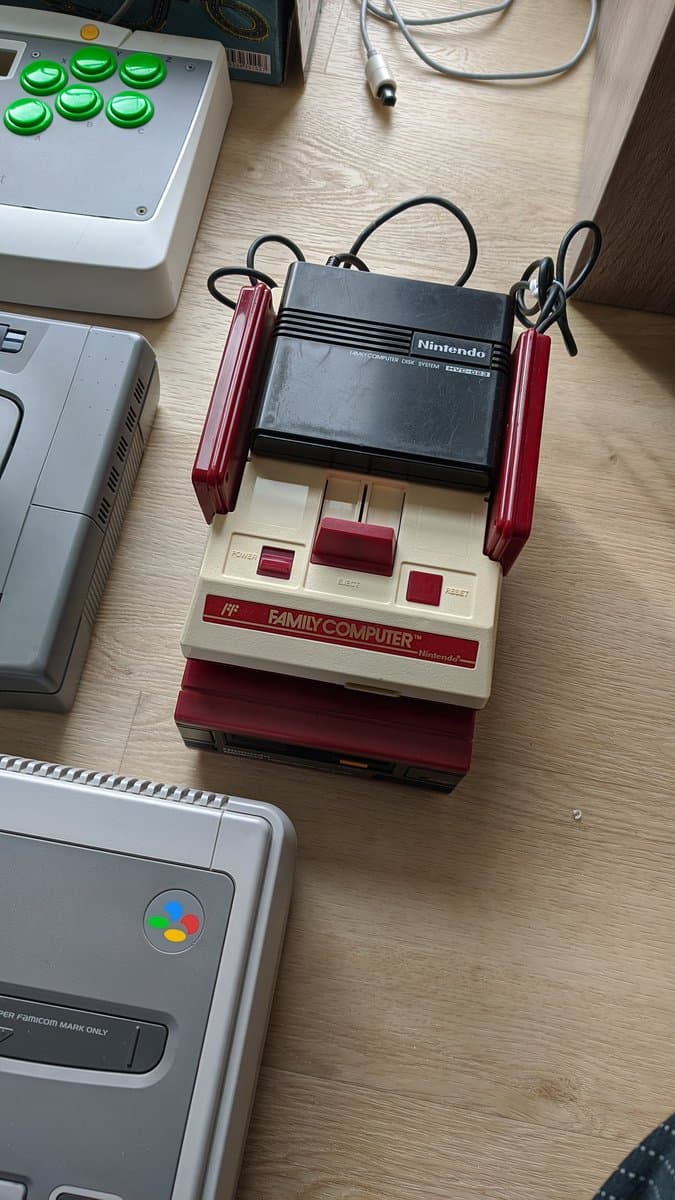 Repaired Nintendo Famicom with red controllers attached, displayed alongside Disk System unit on wooden workbench with other gaming consoles.