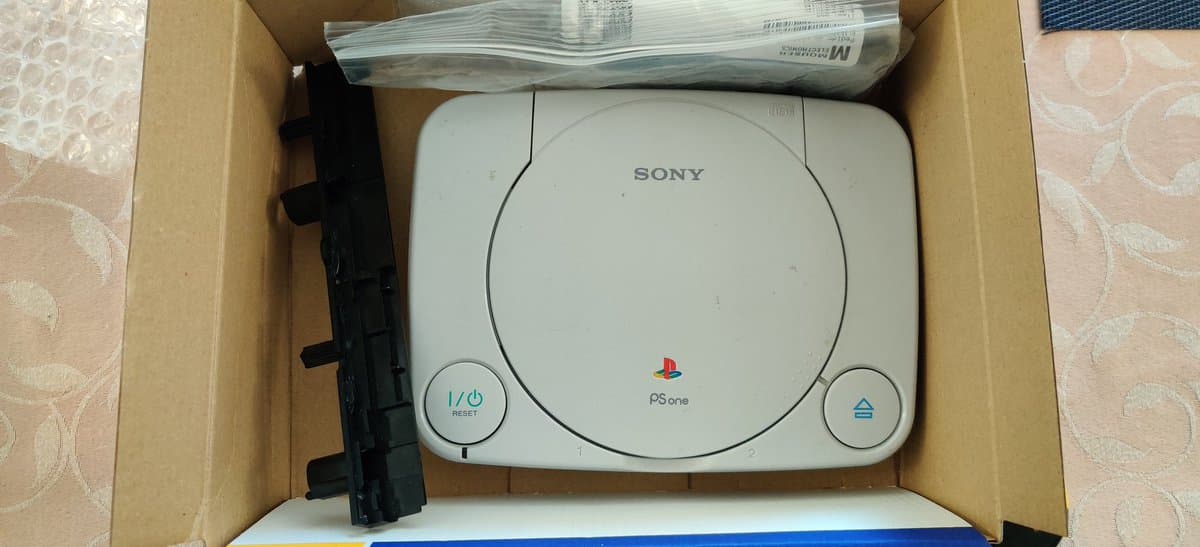Sony PSOne in cardboard box during recap repair, showing white console with controller port and power board removed, documentation visible.