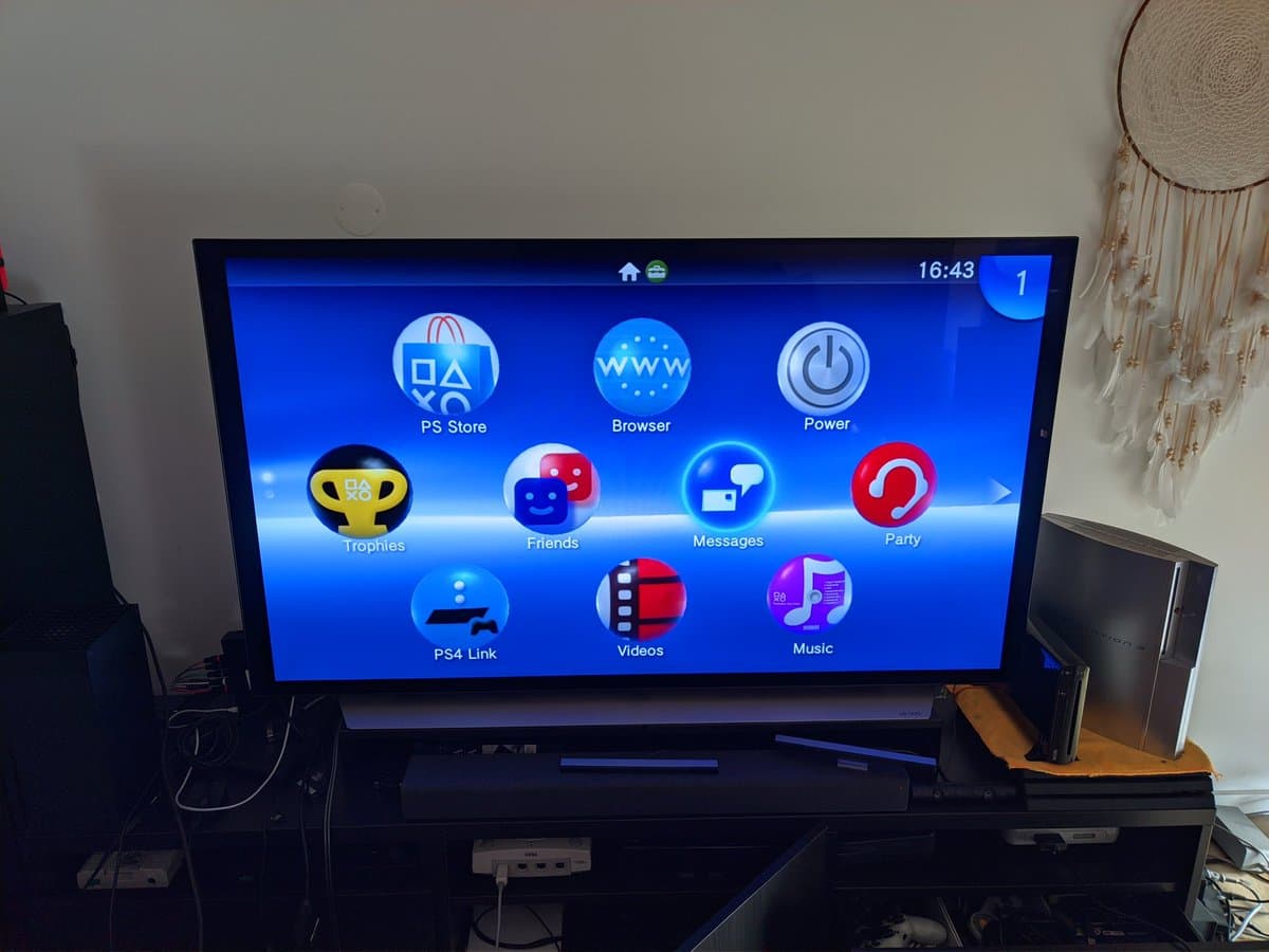 PS Vita TV home menu displayed on TV, showing restored unit with custom firmware installed, positioned on black stand in living room setup.