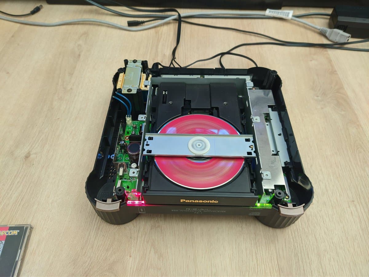 Panasonic FZ-1 3DO console interior with red disc loaded, exposed circuit boards and power supply with blue wiring visible on wooden workbench.