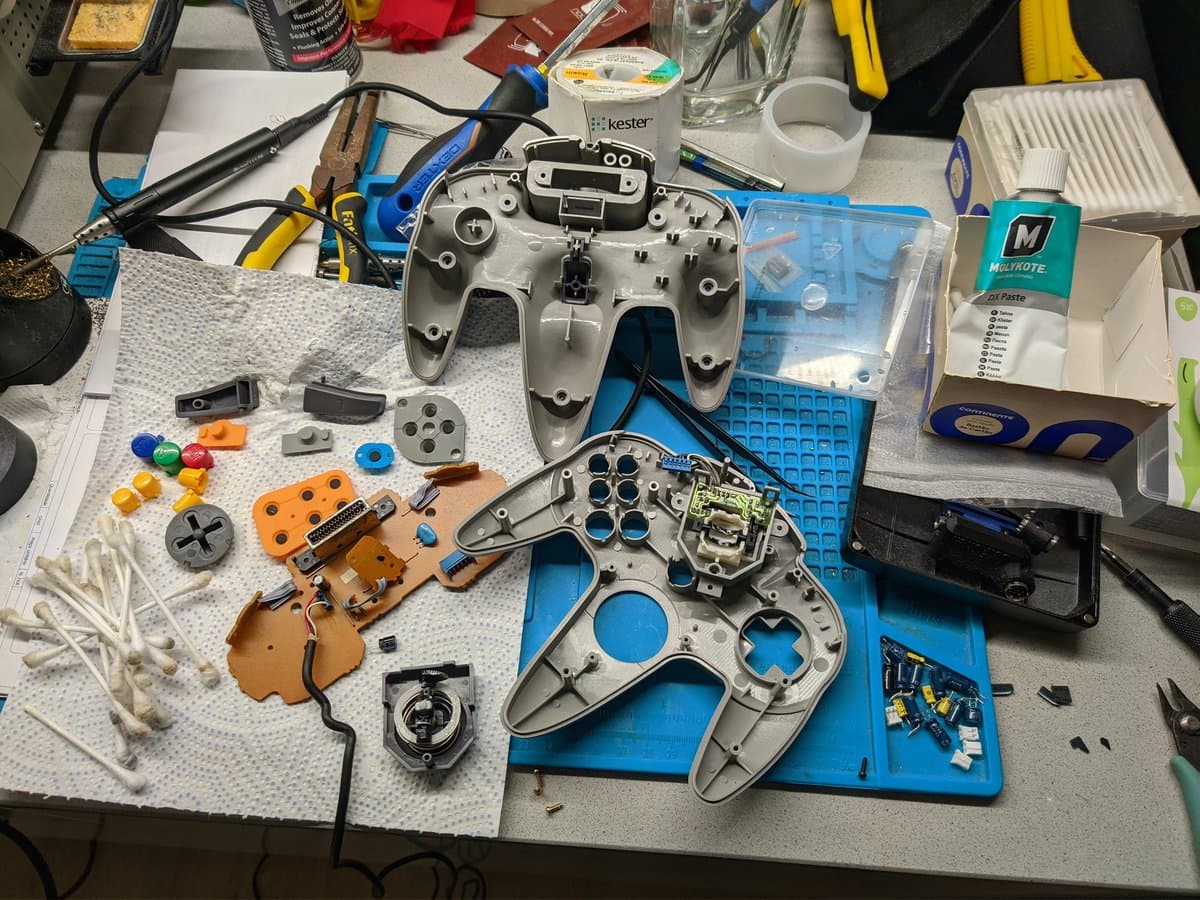 Disassembled N64 controllers and console components on workbench with soldering iron, capacitors, joystick parts, and blue organizer trays during restoration.