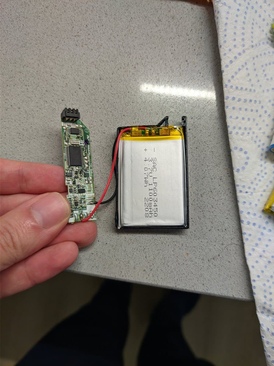 Hand holding PSP-3000 circuit board next to replacement lithium battery with red/black wires in protective casing.