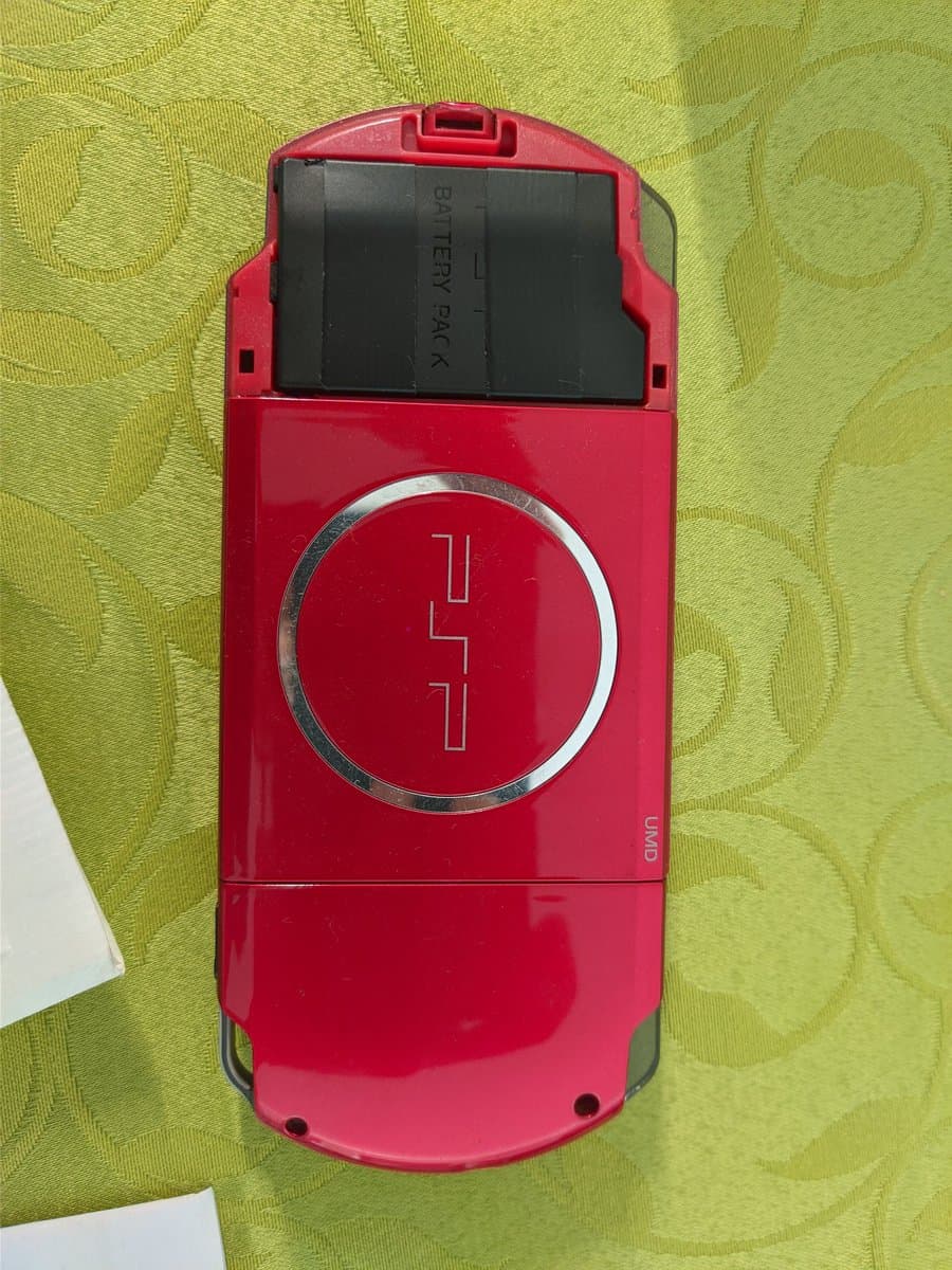 Red PSP-3000 with battery cover open, revealing black battery pack labeled "BATTERY PACK" inside the compartment.