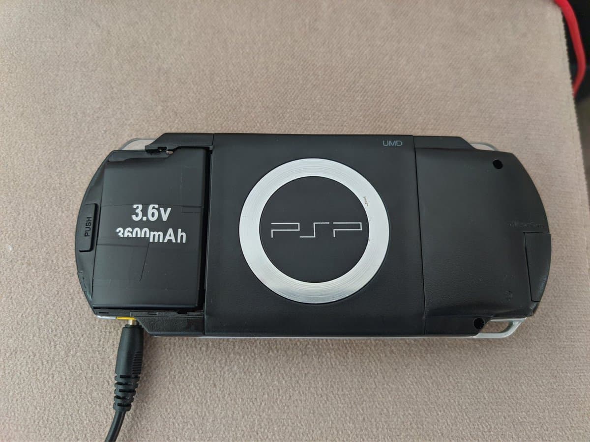 PSP-1000 with open battery door showing 3.6v 3600mAh aftermarket battery case and internal controller board during replacement.