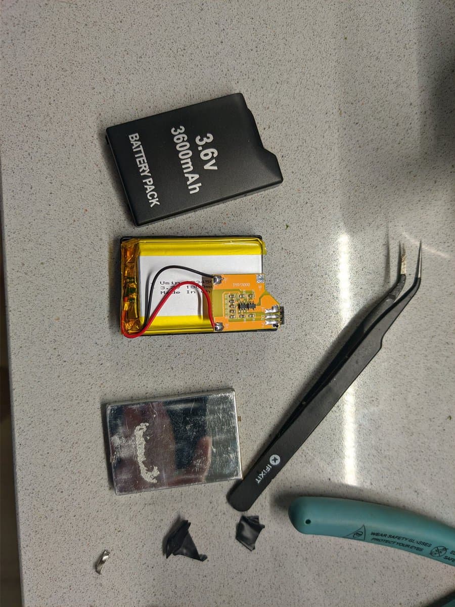 Disassembled PSP-1000 battery components: black case marked 3.6V 3000mAh, yellow lithium cell with circuit board, tweezers, and soldering iron on workbench.