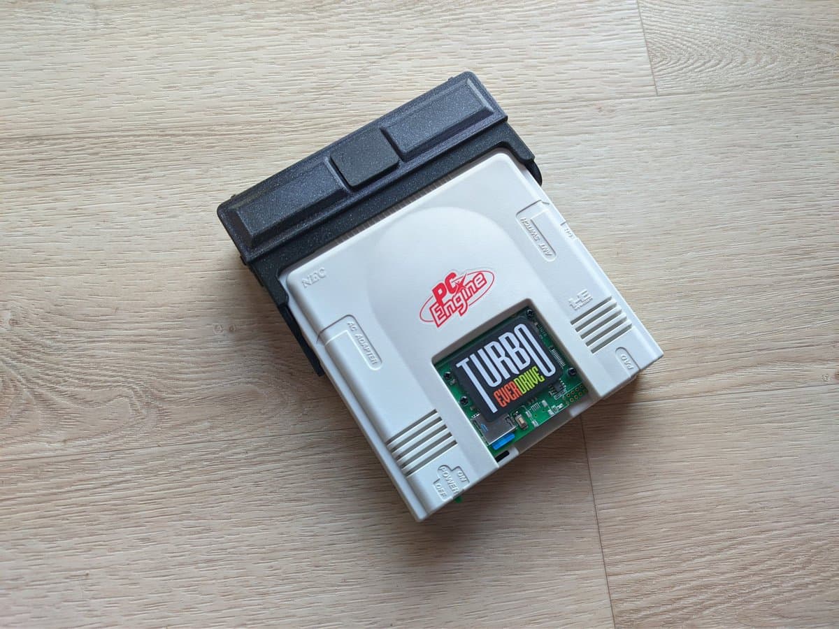 NEC PC Engine with open AV adapter showing internal circuitry and green PCB board labeled "TURBO" on wooden surface.