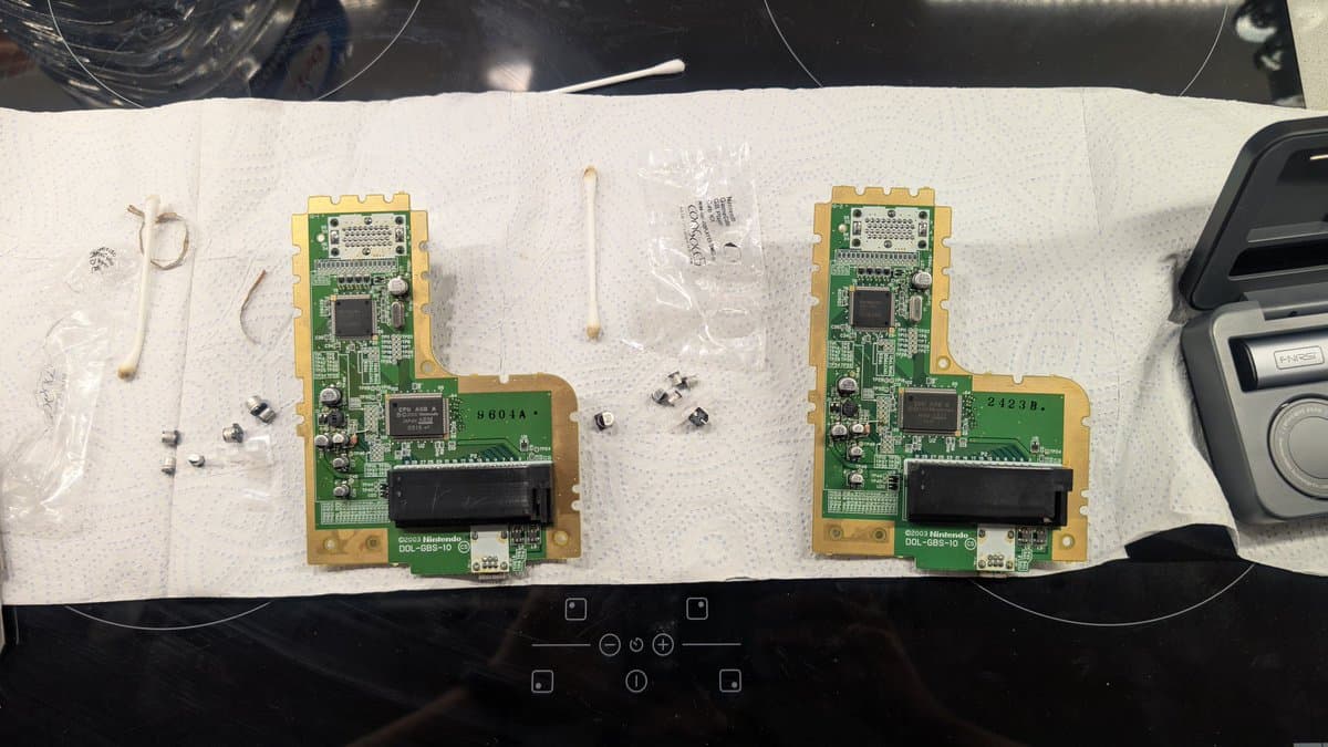 Two Game Boy Player circuit boards during recapping repair, showing removed electrolytic capacitors and desoldering work on white absorbent paper.