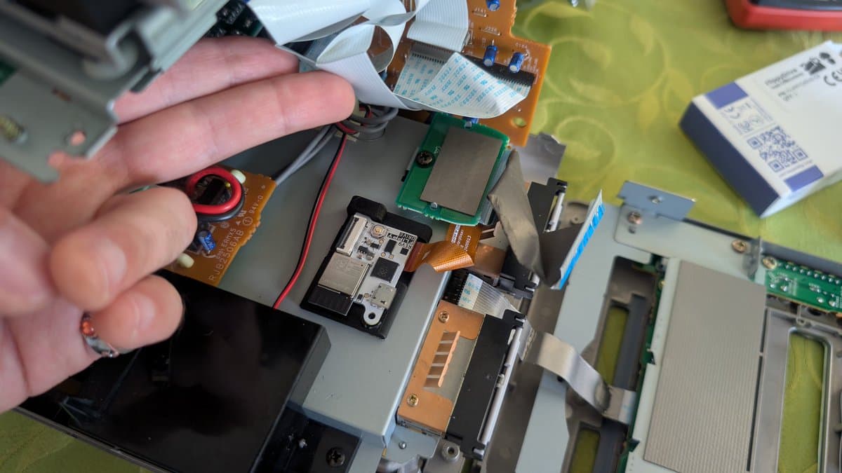 Technician's hand holding flex cable above Panasonic Q optical drive assembly with 3D-printed rails and FlippyDrive installed during restoration.