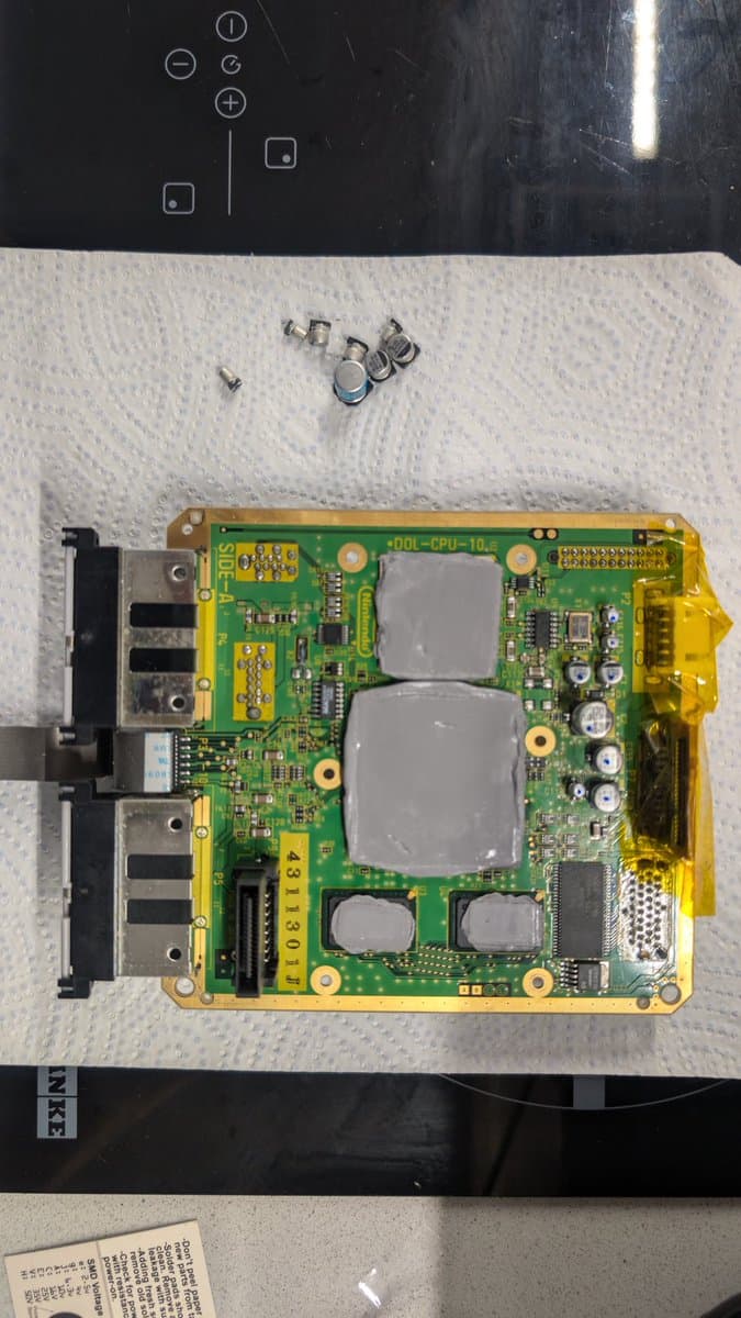 Panasonic Q motherboard during recap, showing two large capacitors removed and small components on tissue paper above the board.