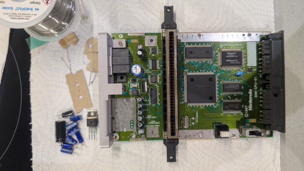 Recapped Super Famicom Jr. motherboard with new regulator, alongside removed capacitors, transistor, and solder spool on workbench.