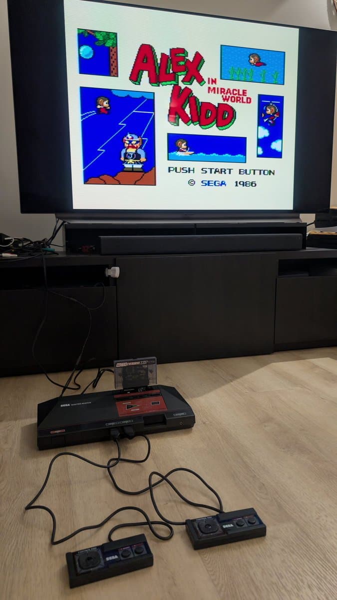 Sega Master System MK-2000 connected to TV displaying Alex Kidd in Miracle World title screen, with two refurbished controllers on wooden surface.