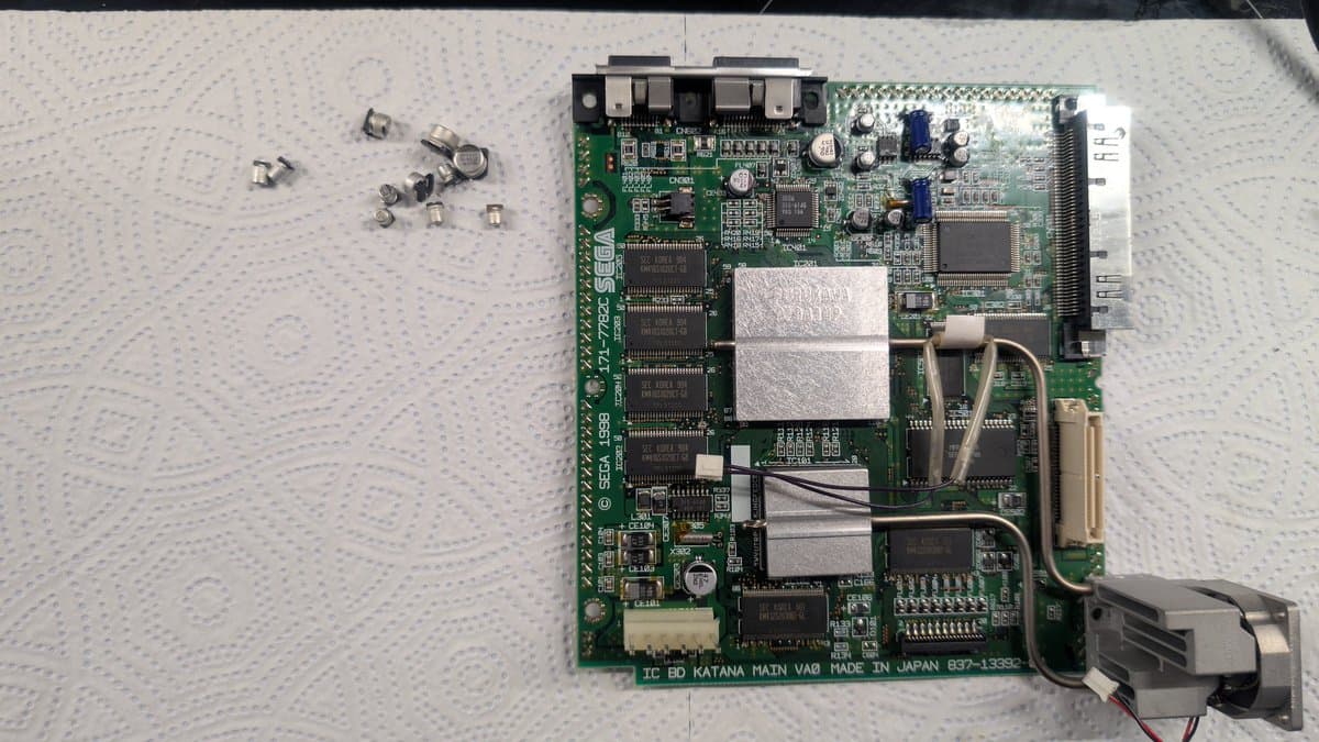 Dreamcast VA0 motherboard with removed capacitors displayed on left, showing refurbishment work in progress on circuit board.