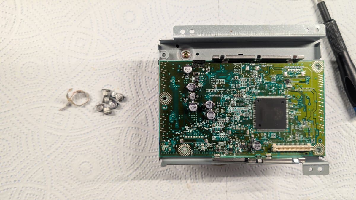 Dreamcast VA0 motherboard with removed capacitors and fuse during refurbishment; metal bracket and screwdriver visible.