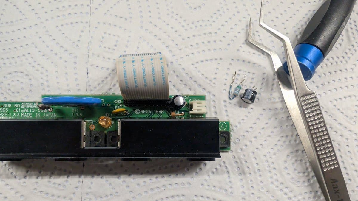 Dreamcast VA0 refurbishment in progress: desoldering old capacitor from motherboard with tweezers and soldering iron, replacement capacitor visible nearby on work mat.
