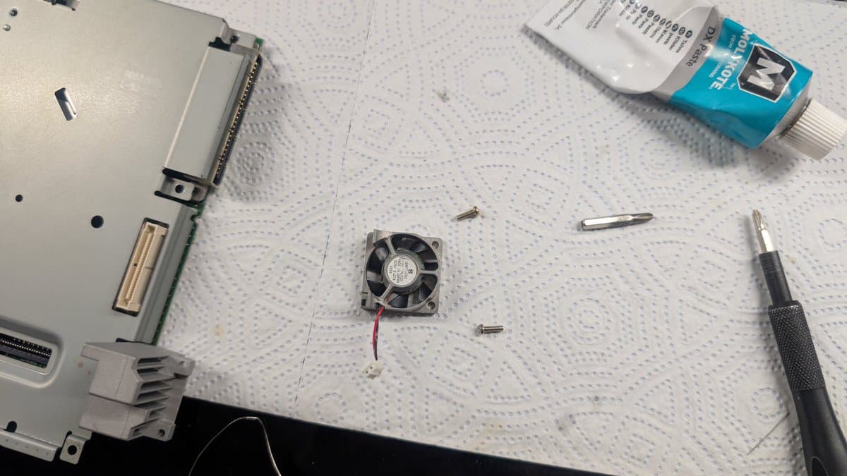 Dreamcast VA0 disassembly: fan with red/black wires, heatsink, screws, thermal paste, screwdriver, and documentation on work mat.