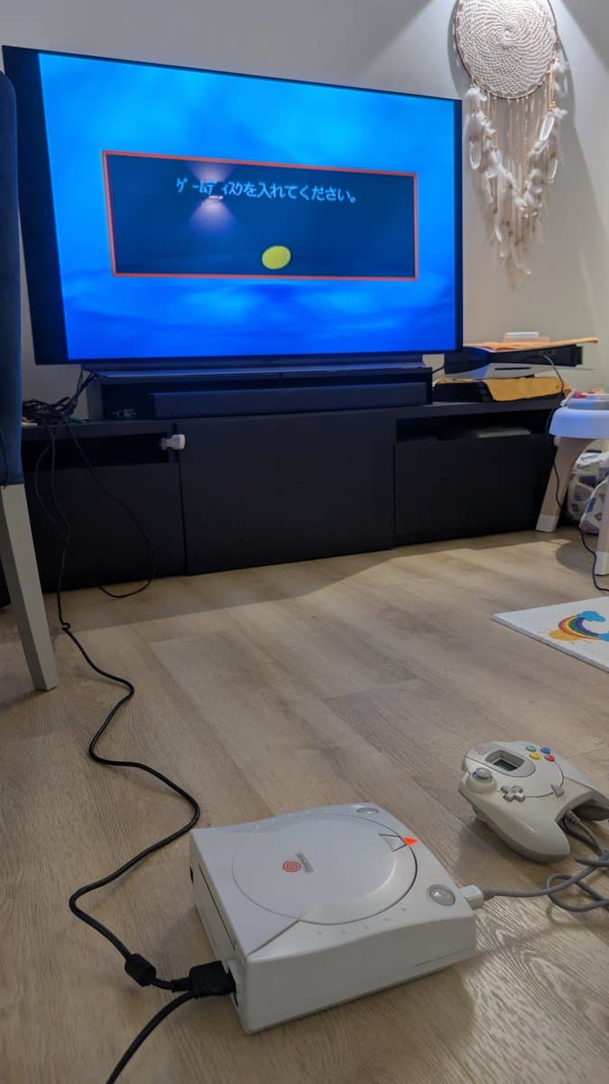 Sega Dreamcast VA0 connected to TV displaying Japanese boot menu with yellow cursor; Dreamcast console and controller visible on wooden floor during testing phase.