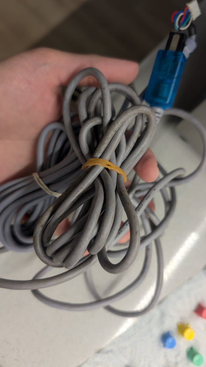 Dreamcast controller cable bundled with gold elastic band, showing gray twisted cord with blue connector end and exposed wires during cleaning process.