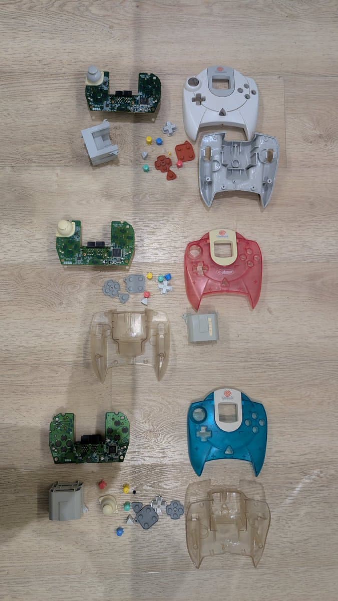 Disassembled Dreamcast controllers in four colors showing circuit boards, shells, buttons, and internal components laid out for cleaning and refurbishment.
