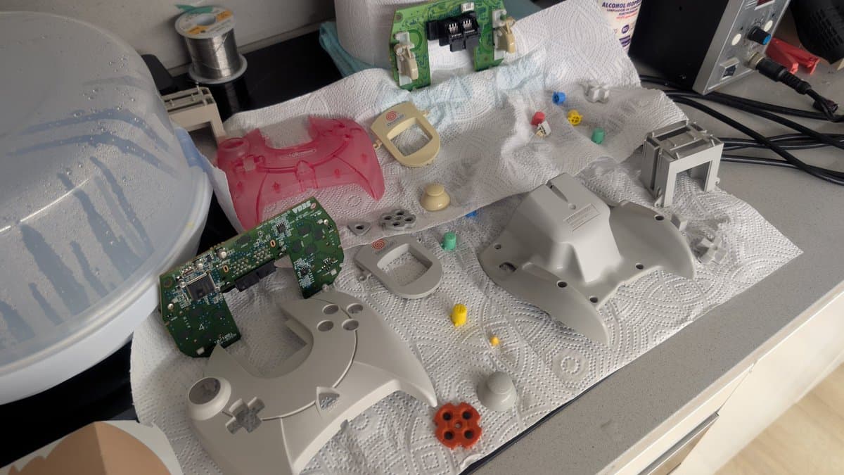 Disassembled Dreamcast VA0 components laid out on white towel: controller shells (pink, white, gray), circuit boards, shell casings, screws, and small colored parts during refurbishment.