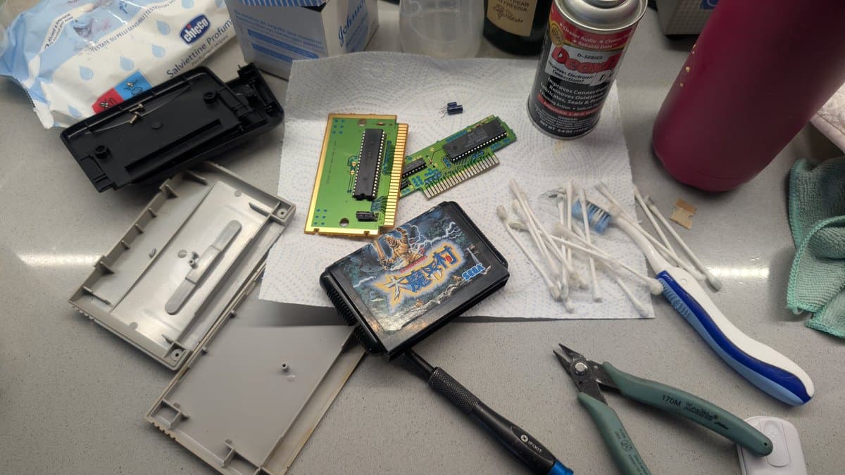 Disassembled game cartridges with PCBs, cleaning supplies, and tools including tweezers and pliers on work surface for maintenance.