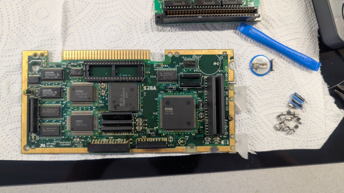 Sega Mega-CD 1 PCB during recap, showing multiple leaking capacitors removed, with soldering iron and desoldered components on white work mat.