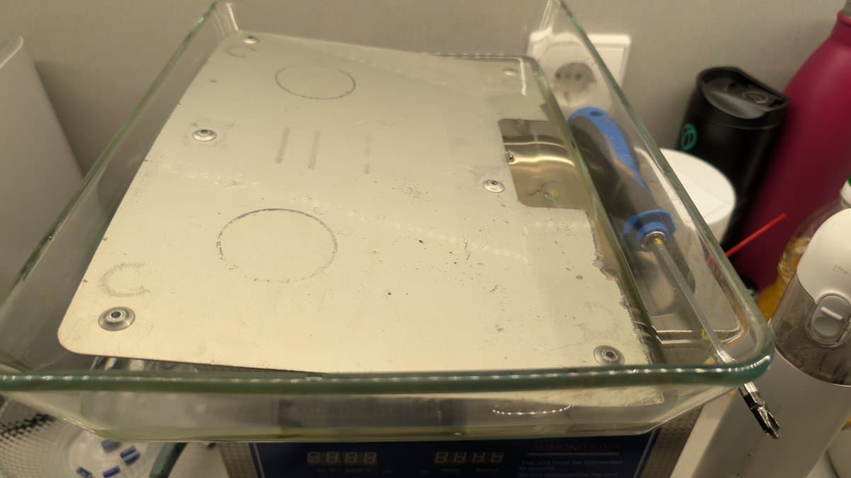 Dreamcast VA0 motherboard and metal shielding plates soaking in Evaporust solution within clear container for rust removal during refurbishment.