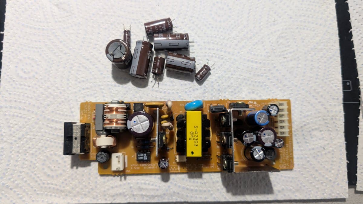 Dreamcast VA0 PSU board with recapped electrolytic capacitors; loose old capacitors displayed above the repaired power supply unit.