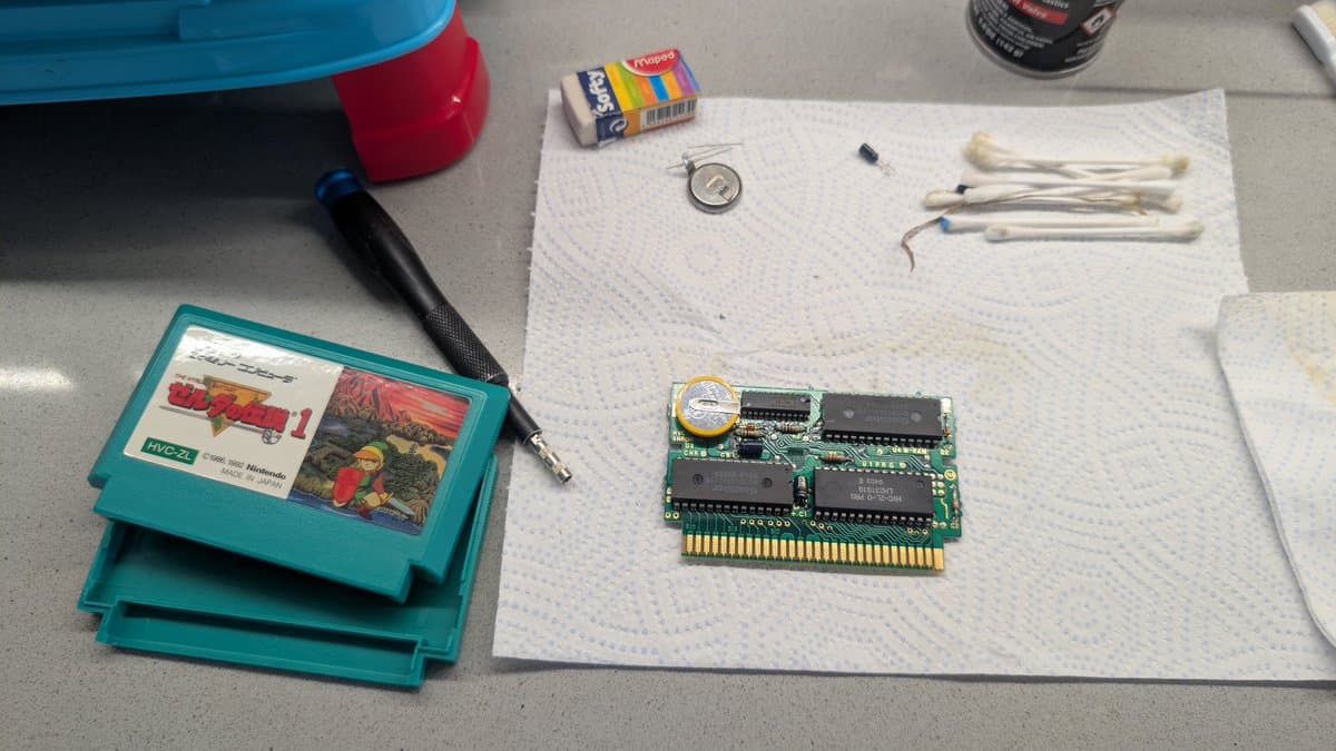Disassembled NES cartridge PCB with new capacitors and coin cell battery holder installed; soldering iron and tools nearby on work mat.