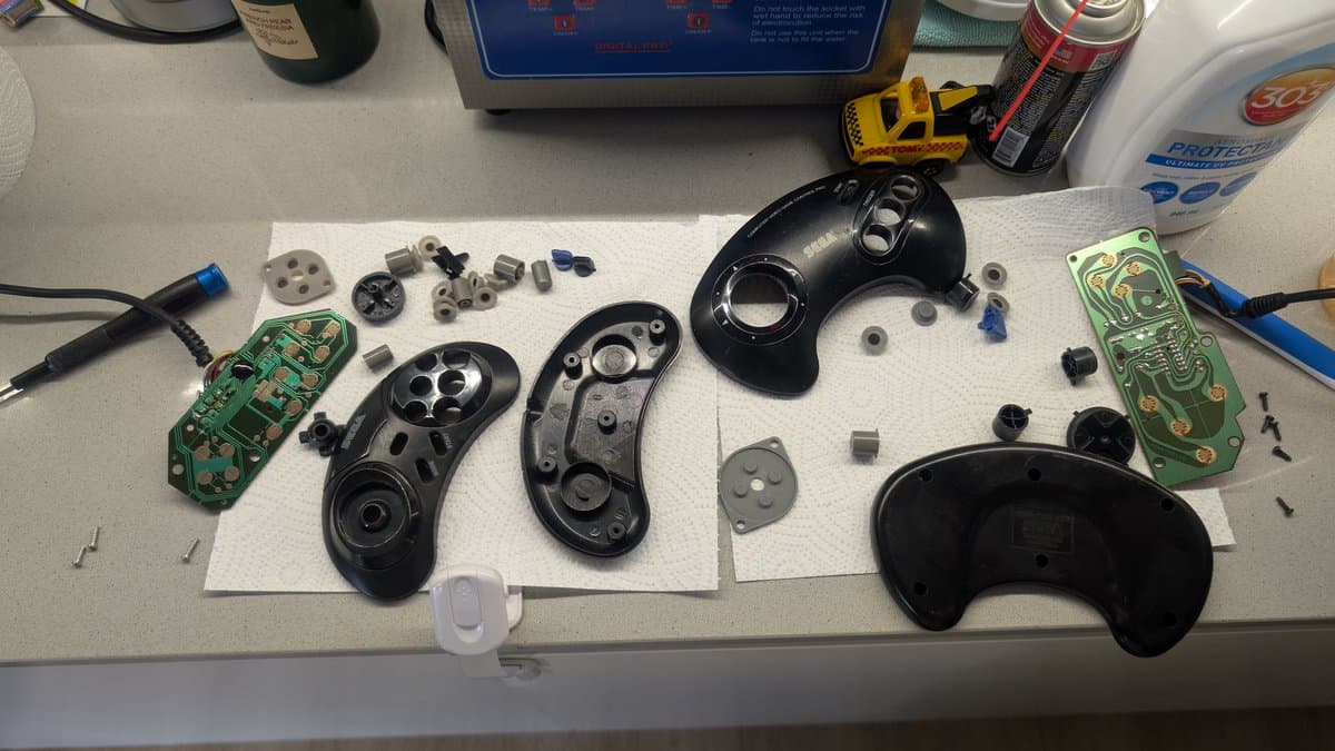 Disassembled Mega Drive controllers and PCBs laid out on workbench during cleaning and inspection, with small components and hardware organized on paper.