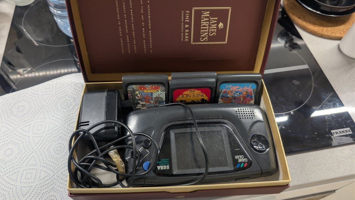 Sega Game Gear in open box with attached transformer inside, power cable, and three game cartridges (Donkey Kong, Lion King, Summer Blast) visible above.