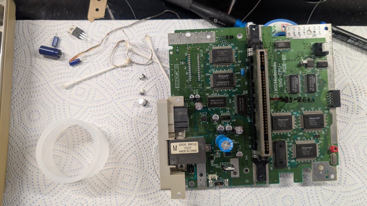 Super Famicom motherboard mid-repair: recapped with new capacitors, voltage regulator replaced, some SMD components remaining; soldering iron and loose components visible on workbench.