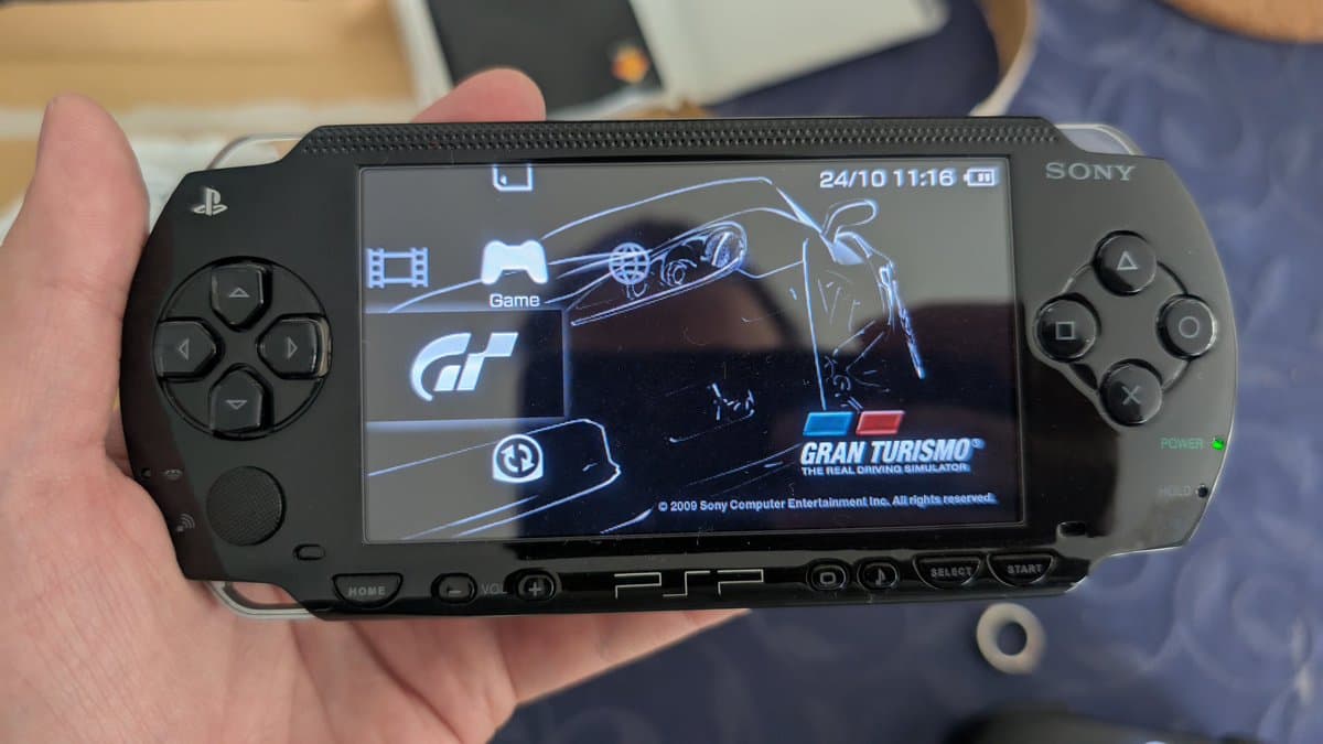 PSP displaying Gran Turismo game menu, visible crack on screen glass, held in hand showing damage from purchase listed as "good condition.