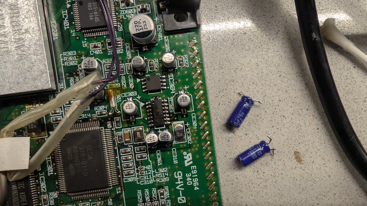 Two blue SMD capacitors removed from Dreamcast motherboard, placed next to circuit board during refurbishment work.