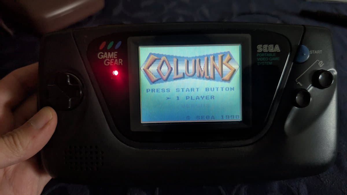 Sega Game Gear displaying Columns title screen with lit power LED, held in hand during ceramic capacitor replacement repair.