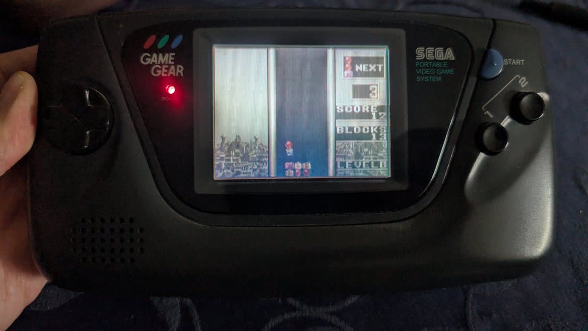Sega Game Gear displaying menu screen with three game options visible, held in hands during ceramic capacitor replacement repair.