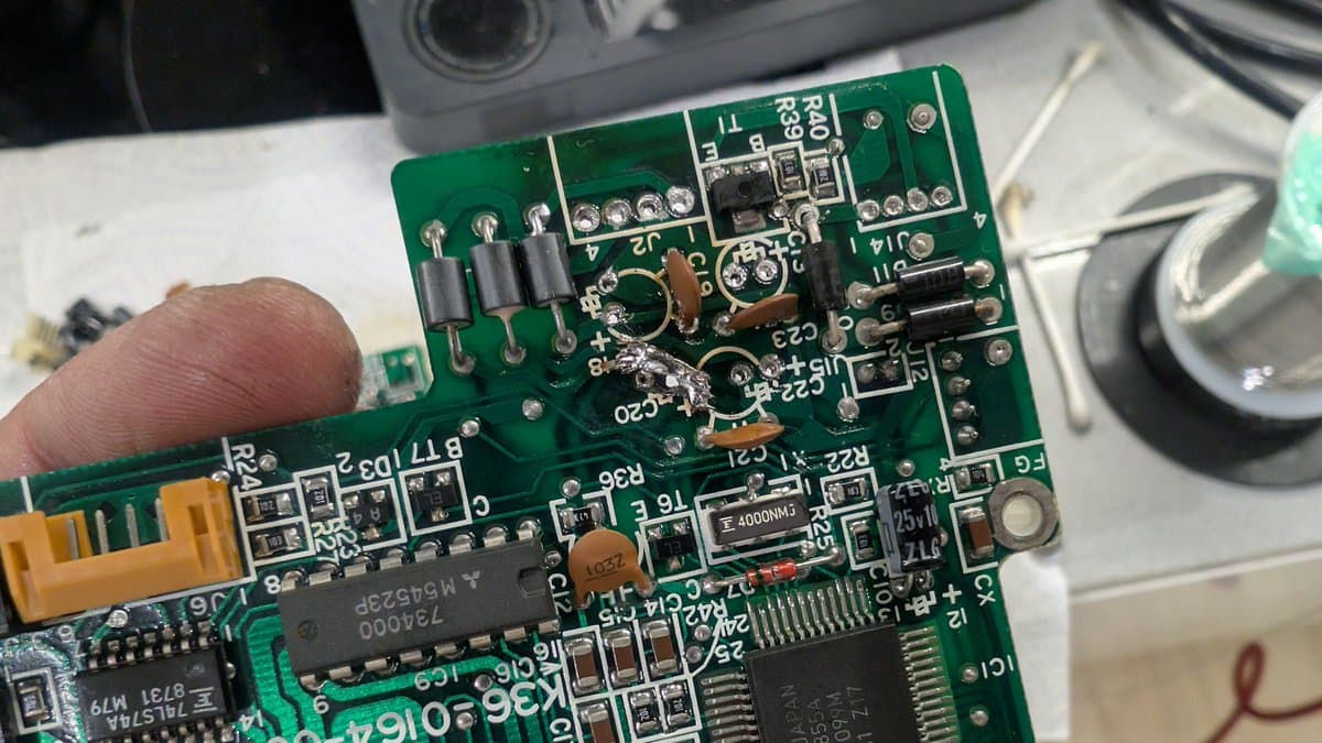 Finger pointing at repaired trace on Sharp X68000 mainboard; brown resistor and solder joints visible near damage area.