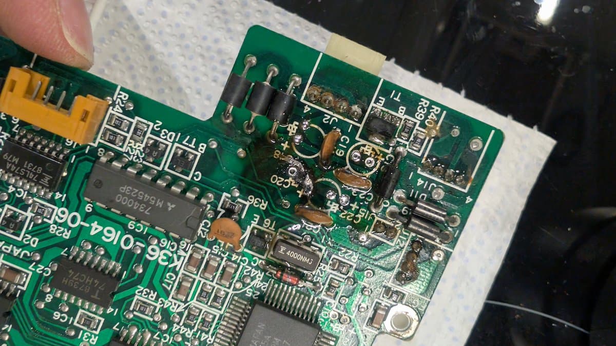 Closeup of Sharp X68000 circuit board showing repaired trace, recapped components, and oxidized solder joints being examined during restoration work.