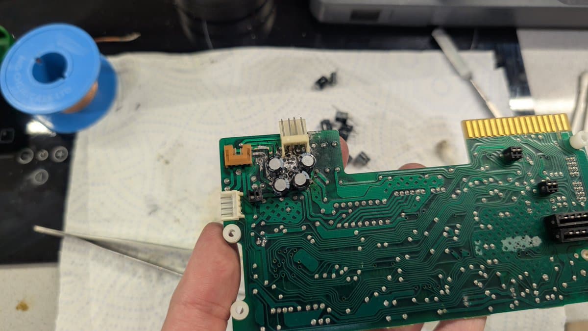 Hand holding Sharp X68000 PCB showing recapped power section with new capacitors, work-in-progress repair on white mat.
