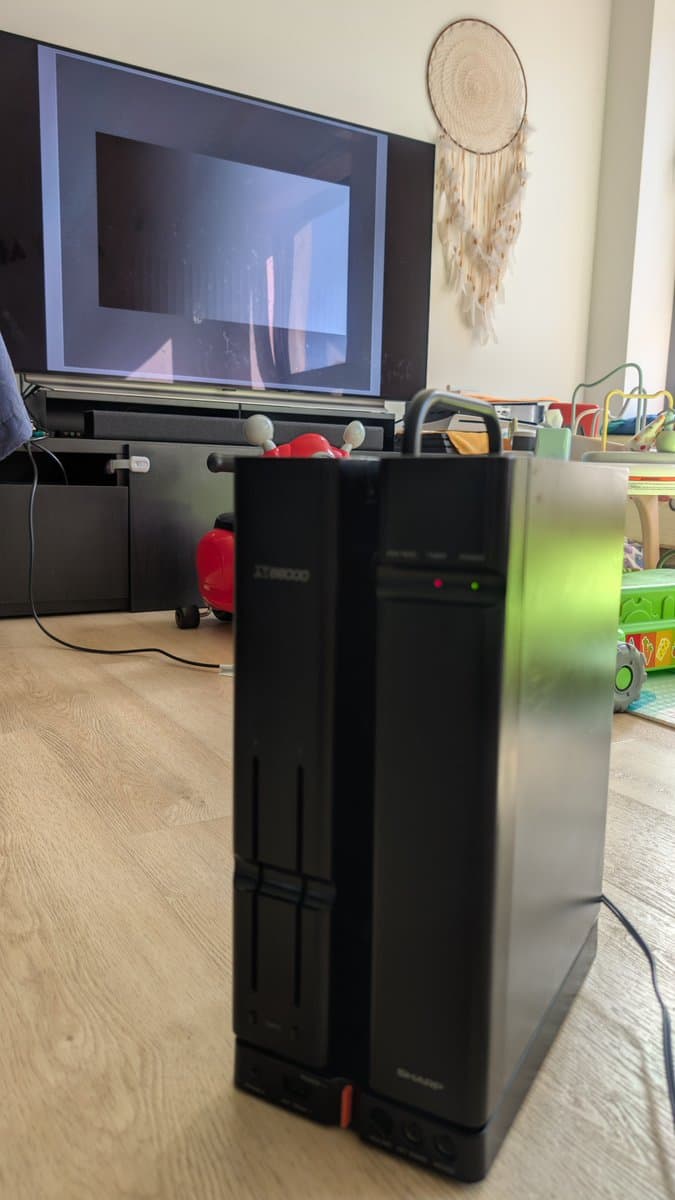 Black Sharp X68000 tower on wooden floor displaying no video output on connected monitor despite power-on state.