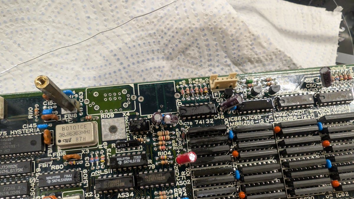 Close-up of Sharp X68000 motherboard showing new oscillator installation, cleaned solder joints near battery leak area, and RAM chips with colored capacitors.