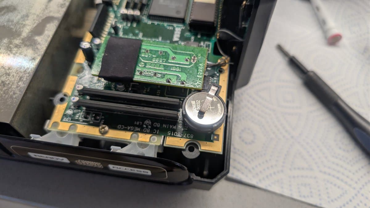 Sega Mega-CD 1 mainboard with new ML2032 coin cell holder installed next to existing battery, showing green PCB and black heatsink.