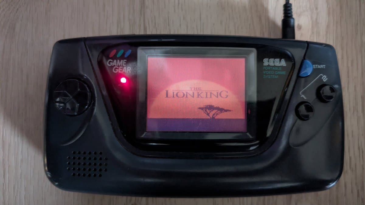 Sega Game Gear displaying Lion King title screen on LCD, power LED lit red, console mounted on wooden surface during repair diagnostic.